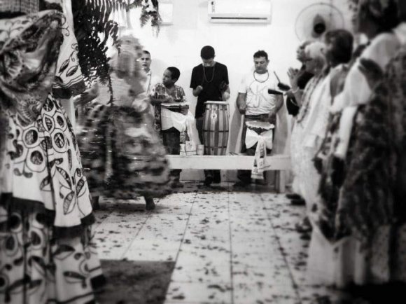 as relações sociais hierárquicas presentes no terreiro, como esclarece Iyá Ejité, o atabaque reverencia um sacerdote ou sacerdotisa e de semelhante forma é reverenciado por uma autoridade da religião ou mesmo por uma divindade.Fonte: Yasmin Estrela, 2019Desta forma, a prática musical nos terreiros de Candomblé é um elemento simbólico propulsor das ações sociais presente no espaço. De acordo com Rosendahl (2012), existe uma dinâmica a qual o homem religioso age sobre o seu ambiente e qualifica-o com formas espaciais diretamente ligadas às suas necessidades. "São marcas simbólicas que respondem aos desejos do devoto em suas práticas espaciais" (Ibid.: 25). As práticas espaciais religiosas contribuem para a criação de um território que possibilita o fortalecimento das experiências religiosas coletivas e individuais garantindo e estabelecendo o domínio dos grupos religiosos no espaço.VI. "Triângulo de Energias" Como Símbolo Hierocósmico no EspaçoAs forças simbólicas responsáveis pela manutenção e construção do espaço sagrado, possuem uma organização à qual obedece à lógica do sagrado (ROSENDAHL, 2014). À esta lógica está presente uma materialidade em que se atribui um valor simbólico, visto que "a modelização simbólica dos objetos espaciais tem, como suporte anterior, os parâmetros da consciência mítica" (GIL FILHO, 2005: 122). A localização dos tambores no espaço reflete a representação cosmogônica iorubá no terreiro revelando um sistema de signos religiosos expressos na paisagem modelados enquanto representações divinas. the Ritual Orchestra: An Ethnography of Musical Instruments in Candomblé Imagem 6: Yemanjá Ogunté reverenciando os tambores após a sua "chegada" ao Ayê.Fonte: Yasmin Estrela, 2016 Imagem 7: Salão Central do Ilè Àsé Iyá Ogunté e ao final estão localizados os atabaques, simultaneamente em direção à porta de entrada. No momento em que fiz o registro desta imagem, em 2016, foi o dia em que realizei a minha primeira entrevista no terreiro. Com o passar dos anos de pesquisa no Ilê, foi possível perceber a profundidade e imanência que os três tambores provocam ao adentrar o salão, onde há uma disposição local dos atabaques em que estão presentes em uma dimensão simbólica da representação, no qual encontram-se as três principais divindades do terreiro: Yemanjá, Ogum e Oyá.